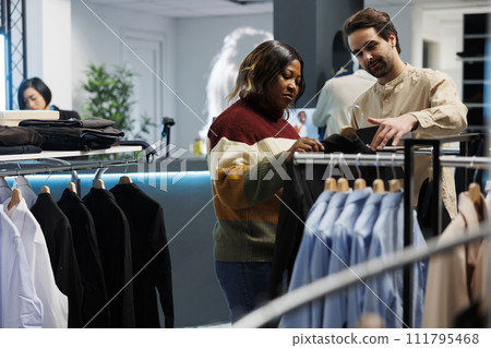 Clothing store customer browsing rack with apparel and talking with assistant. African american woman getting help from boutique employee in choosing trendy outfits while shopping 111795468