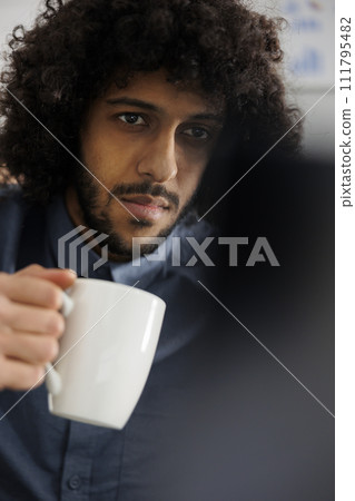 Start up company business employee drinking cofee and doing product management on laptop. Young arab entrepreneur holding tea cup and preparing marketing campaign on computer Start up company business employee drinking cofee and doing product management on laptop. Young arab entrepreneur holding tea cup and preparing marketing campaign on computer 111795482