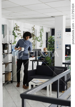 Start up company executive manager working at financial analysis report while standing with laptop near workplace. Arab man drinking coffee and solving business tasks on computer in coworking space 111795484