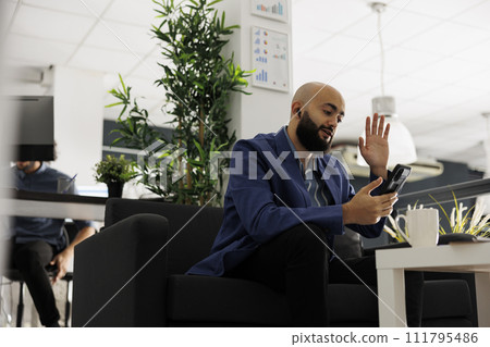 Arab man chatting on smartphone videocall and waving hi in startup business office. Employee greeting team lead in video conference on mobile phone in modern coworking space Arab man chatting on smartphone videocall and waving hi in startup business office. Employee greeting team lead in video conference on mobile phone in modern coworking space 111795486