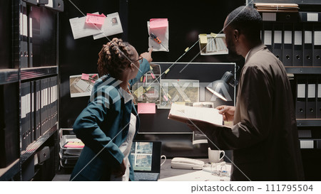 Investigators team talking about suspicious evidence, looking at case files to catch criminal suspect. Two law officers reading witness statements in police archive room, detective work. 111795504