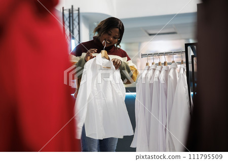 Clothing store buyer trying on shirt while exploring formal apparel for wardrobe. African american woman holding outfit on hanger, examining size, style and fit while shopping in boutique 111795509