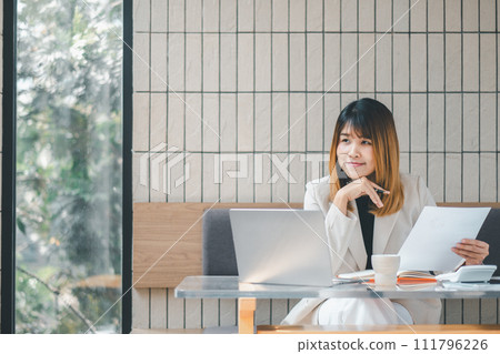 A confident businesswoman assesses documents with a pen in hand, laptop open, in the bright ambiance of a cafe setting. A confident businesswoman assesses documents with a pen in hand, laptop open, in the bright ambiance of a cafe setting. 111796226