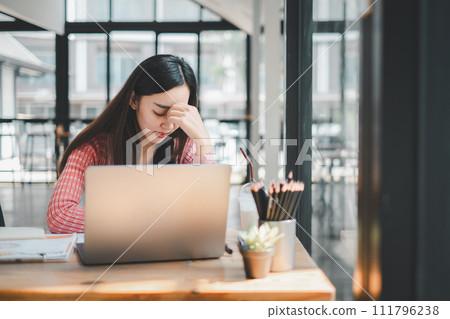 A young woman appears stressed and fatigued while focusing on her laptop screen in a well-lit office environment. A young woman appears stressed and fatigued while focusing on her laptop screen in a well-lit office environment. 111796238