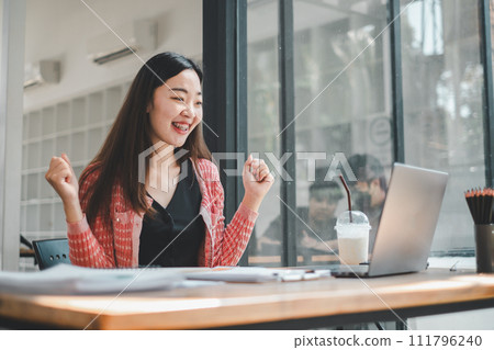 Business analytics concept, An elated young woman celebrates a successful moment at work in front of her laptop in a vibrant office space. 111796240
