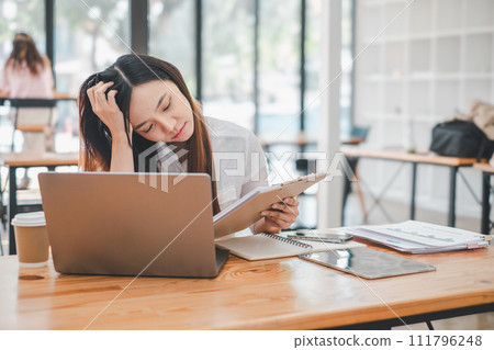 Freelance concept, Worried Asian businesswoman reading documents with a hand on her head, working on a laptop in a busy cafe environment. 111796248