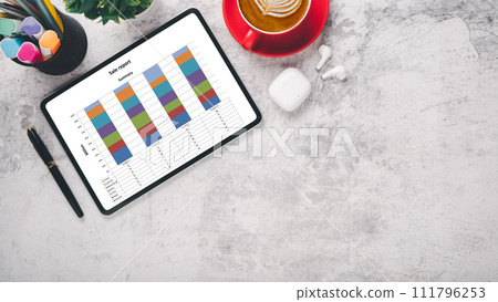 A professional sales report on a tablet with colorful bar graphs, a red coffee cup, and office supplies on a marble surface. A professional sales report on a tablet with colorful bar graphs, a red coffee cup, and office supplies on a marble surface. 111796253