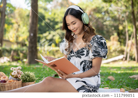 Attractive Asian woman is listening to music on her headphones and reading a book while picnicking. 111799851