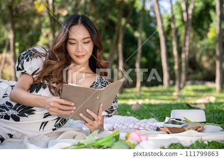 A gorgeous Asian woman is reading a book on a picnic mat while picnicking in a park on a bright day. A gorgeous Asian woman is reading a book on a picnic mat while picnicking in a park on a bright day. 111799863
