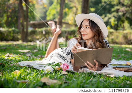 A beautiful and positive Asian woman is lying on a picnic mat and reading a book in a green park. 111799876