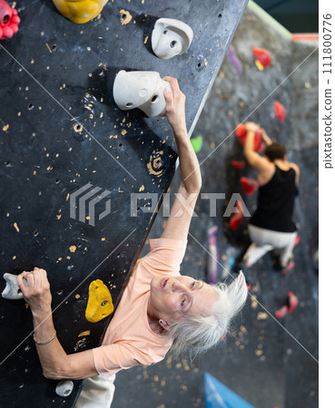 Positive focused mature woman exercising on black climbing wall without safety belts and preparing for summer mountain hiking in game center indoors Positive focused mature woman exercising on black climbing wall without safety belts and preparing for summer mountain hiking in game center indoors 111800776