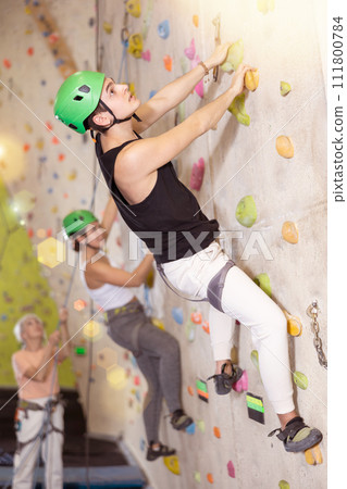 Side view of sportive young male climber moving up, using safety top rope and climbing artificial wall indoors. Extreme sports and bouldering concept Side view of sportive young male climber moving up, using safety top rope and climbing artificial wall indoors. Extreme sports and bouldering concept 111800784