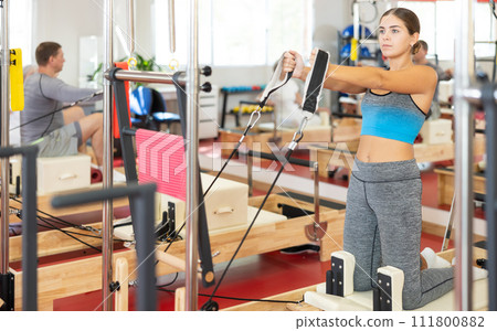 Sports club girl client on reformer with cable and rope performs exercise to stretch muscles of arms 111800882