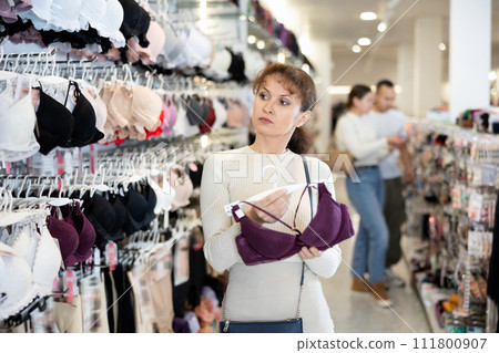 Woman stopped near showcase with lingerie, holds bust bodice hanger in hands and examines product 111800907