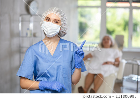 Happy doctor or nurse with mask in uniform with crossed arm, patient behind on couch. 111800993