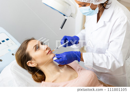Cosmetologist making injections of botulotoxin into lips of young woman 111801105