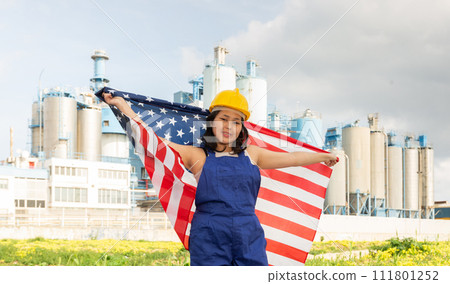 Happy young woman in helmet holding flag of the USA against background of factory Happy young woman in helmet holding flag of the USA against background of factory 111801252