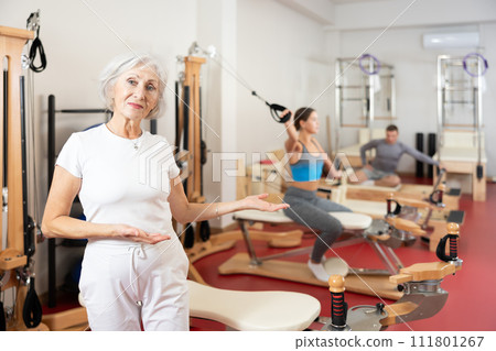 Positive senior woman coach stands in large gym against background of Pilates simulators 111801267