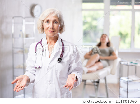 Smiling senior woman doctor in uniform with patient lying behind in clinic. Smiling senior woman doctor in uniform with patient lying behind in clinic. 111801360