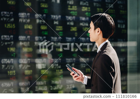 一名身穿西裝的 30 多歲日本男子一邊用智慧型手機進行 NISA 交易,一邊看著公告牌上顯示的即時股價板。 一名身穿西裝的 30 多歲日本男子一邊用智慧型手機進行 NISA 交易,一邊看著公告牌上顯示的即時股價板。 111810166