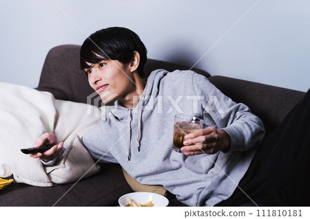 Lazy man in his thirties lying on the sofa and relaxing while drinking alcohol and operating the remote control to change the channel 111810181
