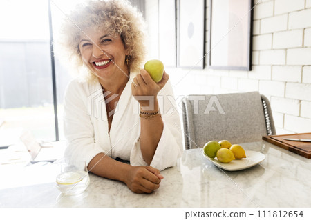 Cheerful Woman in White Robe Holding Apple in Sunny Kitchen Cheerful Woman in White Robe Holding Apple in Sunny Kitchen 111812654
