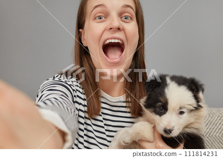 Beautiful young adult cheerful woman taking selfie on smartphone with her dog exclaiming with excitement and happiness keeps mouth widely opened making point of view photo for her network page 111814231