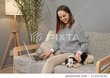 Smiling Caucasian woman wearing striped shirt sitting on sofa with her puppy dog using laptop computer working online and patting her small black and white dog Smiling Caucasian woman wearing striped shirt sitting on sofa with her puppy dog using laptop computer working online and patting her small black and white dog 111814242