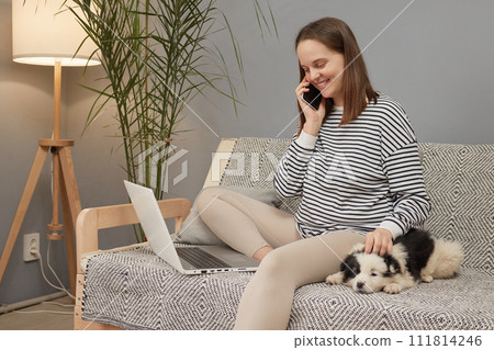 Smiling cheerful Caucasian woman wearing striped shirt sitting on sofa with her puppy dog using laptop computer and talking on smartphone having pleasant conversation with colleague Smiling cheerful Caucasian woman wearing striped shirt sitting on sofa with her puppy dog using laptop computer and talking on smartphone having pleasant conversation with colleague 111814246