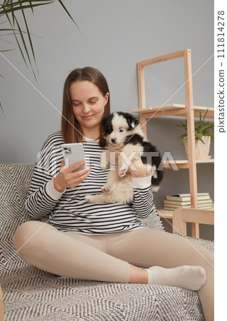 Beautiful young adult woman sitting with cute little dog on couch using mobile phone for online scrolling surfing internet female wearing striped shirt looking at smartphone display Beautiful young adult woman sitting with cute little dog on couch using mobile phone for online scrolling surfing internet female wearing striped shirt looking at smartphone display 111814278