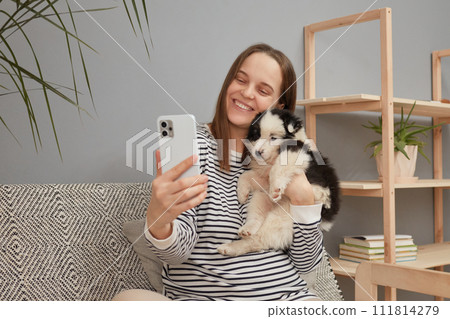 Smiling satisfied young adult woman wearing striped shirt taking selfie on smartphone with her small dog while sitting on sofa at home creating content for her blog 111814279
