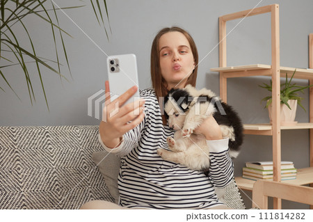 Adorable young adult woman wearing striped shirt taking selfie on smartphone for her blog with her small dog while sitting on sofa at home sending air kissing for phone display 111814282