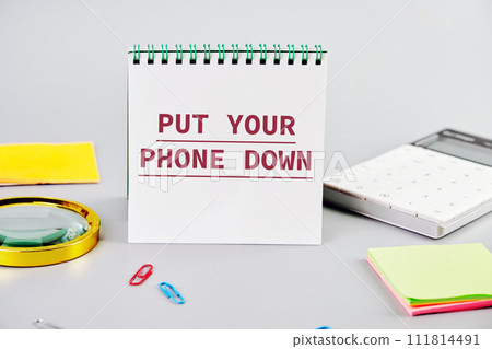 Put Your Phone Down text written on a blank sheet of notepad next to office supplies on a gray background. Business concept Put Your Phone Down text written on a blank sheet of notepad next to office supplies on a gray background. Business concept 111814491