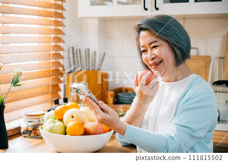 In the kitchen, an old woman, a grandmother, savors breakfast. She's smiling, using her smartphone, and munching on a red apple. A portrayal of technology, happiness, and care at home. In the kitchen, an old woman, a grandmother, savors breakfast. She's smiling, using her smartphone, and munching on a red apple. A portrayal of technology, happiness, and care at home. 111815502