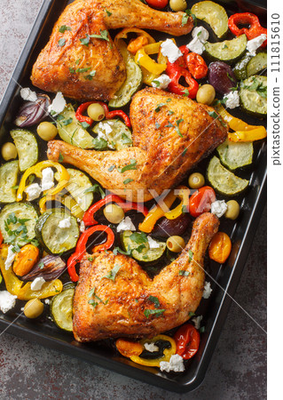 Mediterranean diet baked chicken legs with zucchini, olives, onions, tomatoes, peppers and feta cheese close-up on a baking sheet on the table. Vertical top view 111815610