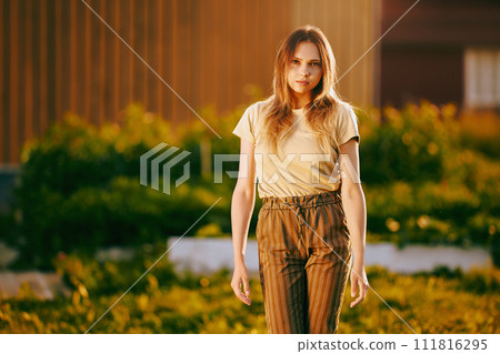 Knee-length portrait of young slender pretty woman in striped trousers and T-shirt at sunset. 111816295