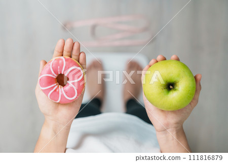 woman on weight scale and hand hold green Apple and donut, choose between fruit is Healthy and sweet is Unhealthy junk food. Dieting control, Weight loss, Obesity, eating lifestyle and nutrition 111816879
