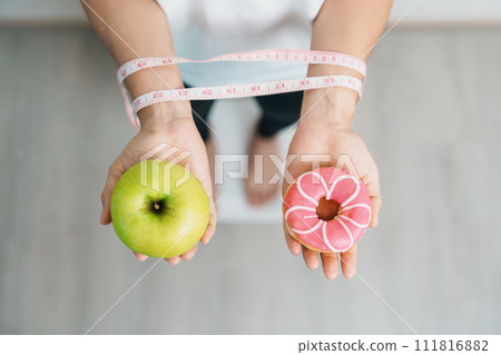 woman on weight scale and hand hold green Apple and donut, choose between fruit is Healthy and sweet is Unhealthy junk food. Dieting control, Weight loss, Obesity, eating lifestyle and nutrition 111816882