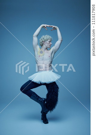 Shirtless man in tutu and boots posing in ballet stance against grey studio background, embodying strength and grace. 111817908