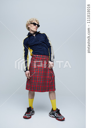 Full-length portrait of young curly haired man posing in Scottish kilt and sportwear against grey studio background. 111818016