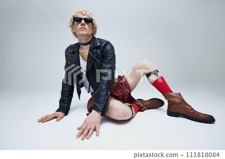 Funny man in traditional Scottish outfit and modern punk leather jacket posing lying on floor against grey studio background. Funny man in traditional Scottish outfit and modern punk leather jacket posing lying on floor against grey studio background. 111818084