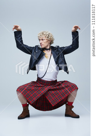 Man in kilt and leather jacket posing with raised arms, embodying mix of traditional and punk style against grey studio background. 111818117
