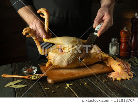 A cook cuts a raw rooster with a knife in the kitchen of a public house. Concept of preparing a chicken dish for lunch on a dark background 111818704