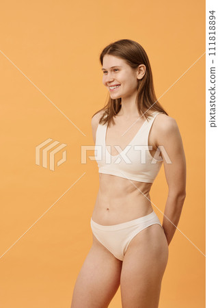 Vertical medium long shot of joyful young Caucasian woman with no makeup wearing minimalistic lingerie standing in studio, copy space 111818894