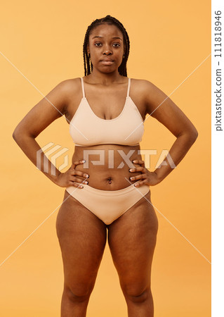 Vertical medium long studio portrait of confident young Black woman wearing minimalistic lingerie posing for camera with hands on hips Vertical medium long studio portrait of confident young Black woman wearing minimalistic lingerie posing for camera with hands on hips 111818946
