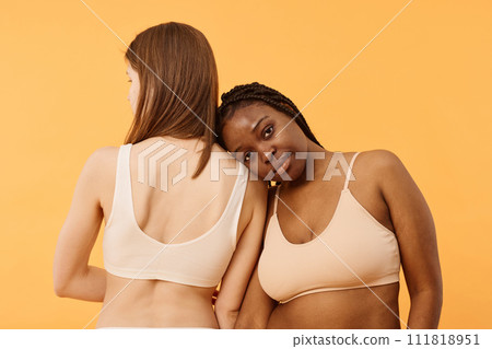 Medium studio shot of two young ethnically diverse women wearing comfortable bralettes posing for camera, warm apricot background 111818951