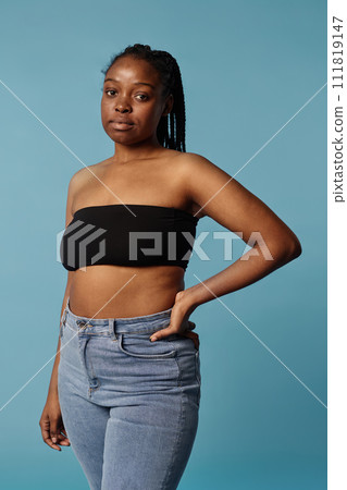 Vertical medium long portrait of young African American woman wearing bandeau top and jeans posing for camera in studio 111819147