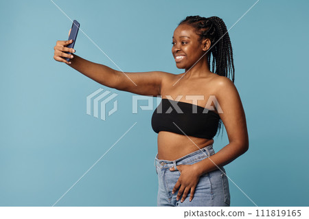 Medium studio portrait of cheerful young African American woman wearing bandeau top and jeans taking selfie on smartphone Medium studio portrait of cheerful young African American woman wearing bandeau top and jeans taking selfie on smartphone 111819165