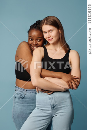 Vertical medium long studio shot of two modern young Black and Caucasian women posing for camera hugging 111819166