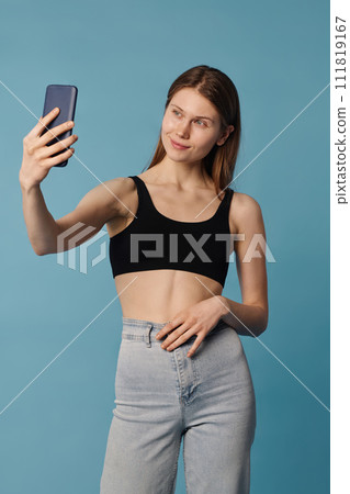 Vertical medium long studio shot of young Caucasian woman wearing sports bra and jeans taking selfie on smartphone 111819167